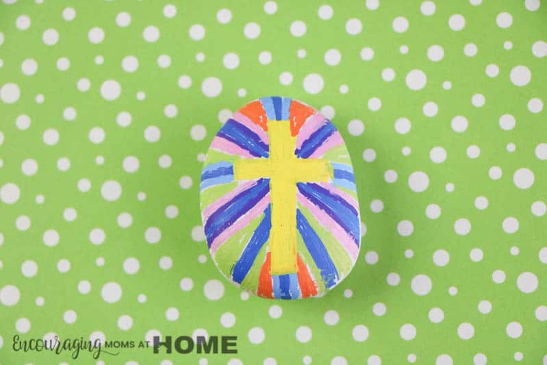 cross painted rock with yellow color on green background.